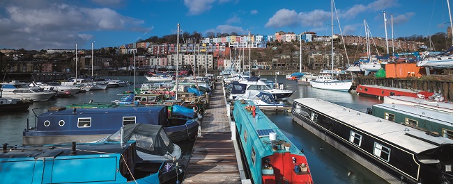Colourful boats on the harbourside, with colourful rows of houses in the distance on a blue sunny day. 